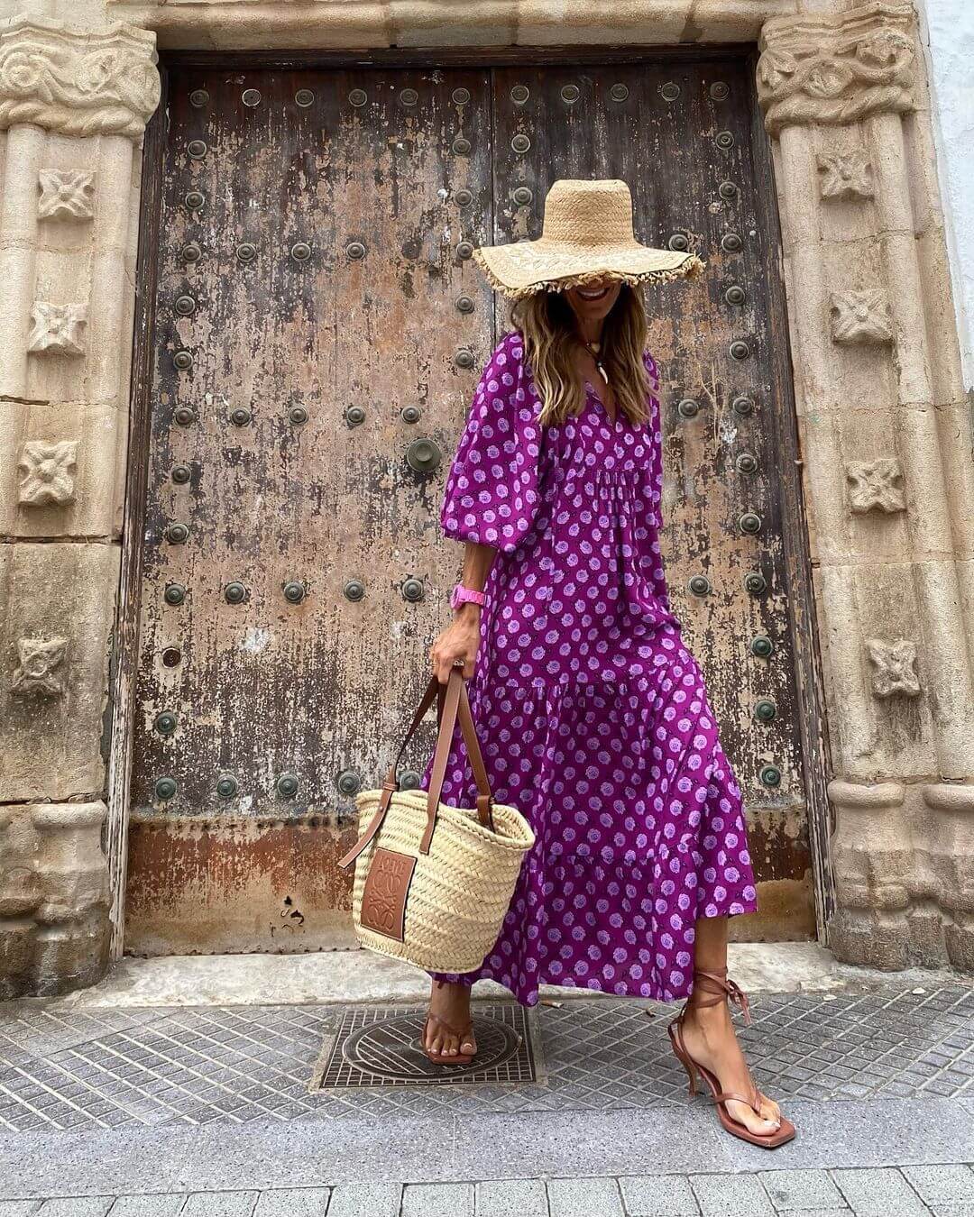 woman wearing purple British style vintage bohemian floral long dress with wide sleeves and straw hat carrying woven tote bag standing in front of ornate wooden door