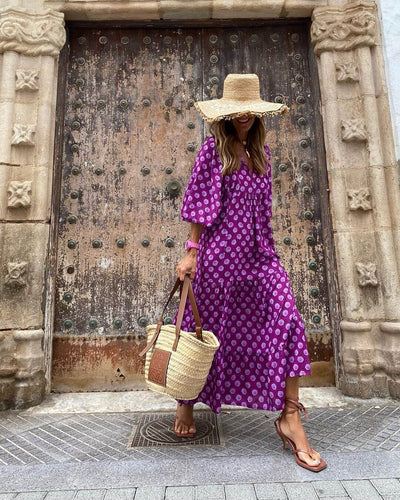 woman wearing purple British style vintage bohemian floral long dress with wide sleeves and straw hat carrying woven tote bag standing in front of ornate wooden door