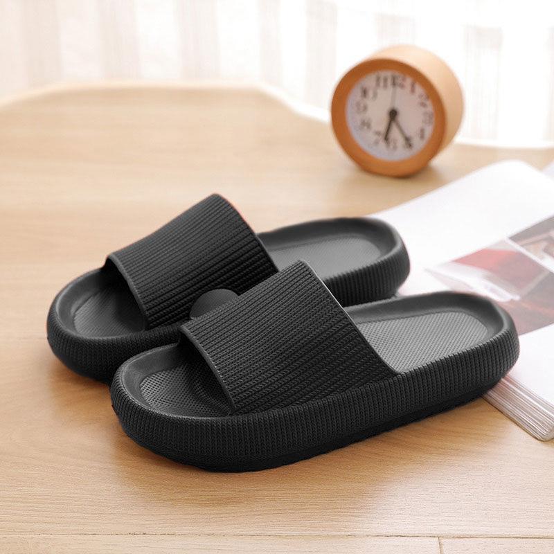Black bathroom sandals on wooden floor with clock in background.